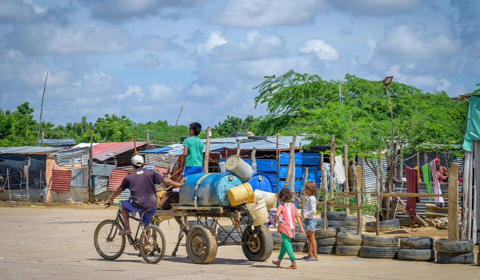 A child rides on top of a cart carrying buckets, while two children walk at the back and another rides their bicycle beside it. (Colombia, 2023).