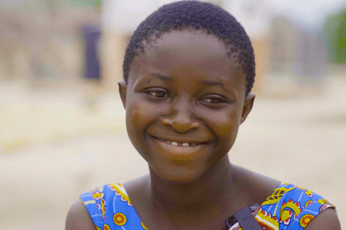 A young girl in a blue patterned shirt, smiles. (Ghana, 2024).