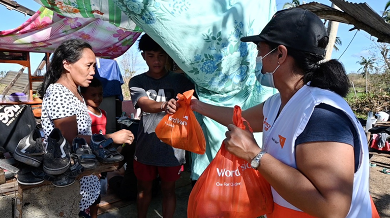 A lady in a mask gives bags of food to another lady in a makeshift shlter