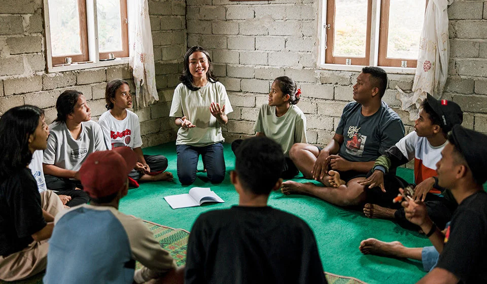A girl is speaking to a group of adolescent girls and boys gathered in a room.